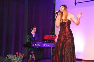 Zdjęcie kobiety śpiewającej na scenie podczas koncertu, z tyłu niej mężczyzna grający na pianinie