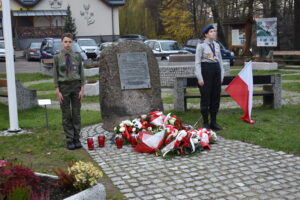 Zdjęcie kwiatów pod kamieniem upamiętniające Święto Niepodległości, obok kamienia jest 2 harcerzy