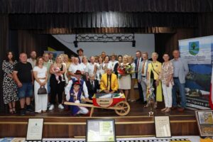 Zdjęcie przedstawia grupę osób na scenie, wśród nich jest Dyrektor GOKSiR i Wójt Gminy Lipusz, część ubrana jest w kaszubskie stroje
