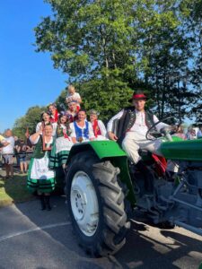 Zdjęcie przedstawia mężczyzne siedzącego w ciągniku ubranego w strój kaszubski, z tyłu niego jest pare kobiet również ubranych w strój kaszubski