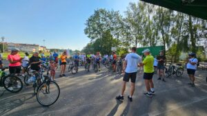 Zdjęcie przedstawia uczestników Rajdu Rowerowego i Dyrektora GOKSiR, mówiącego przez mikrofon, w tle widać szkołe i stadion