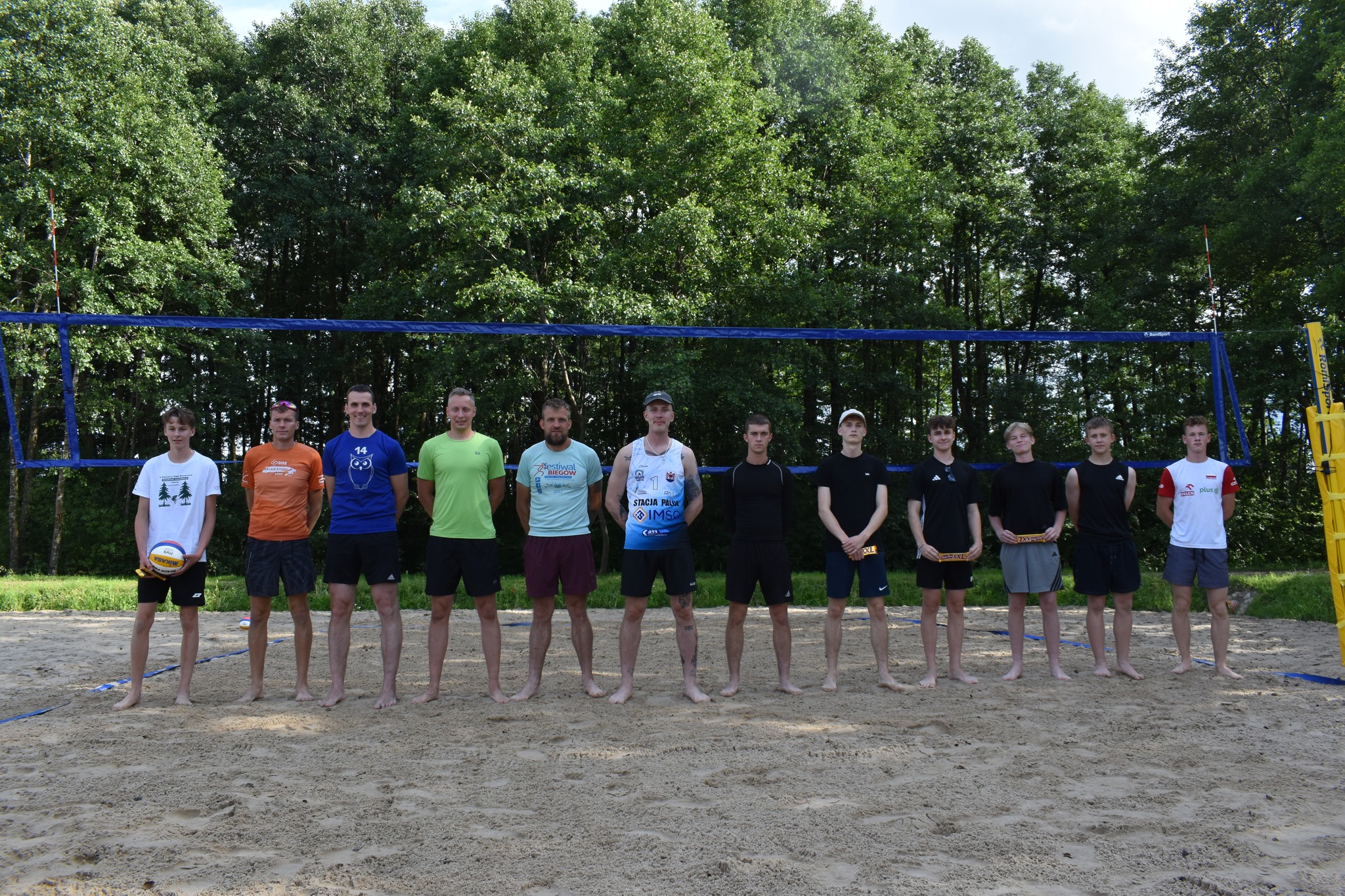 Na zdjęciu widać 12 zawodników siatkówki plażowej, którzy uczestniczyli w 3 turnieju siatkówki plażowej z cyklu Grand Prix Lipusza 2025, w tle siatka i zielone drzewa