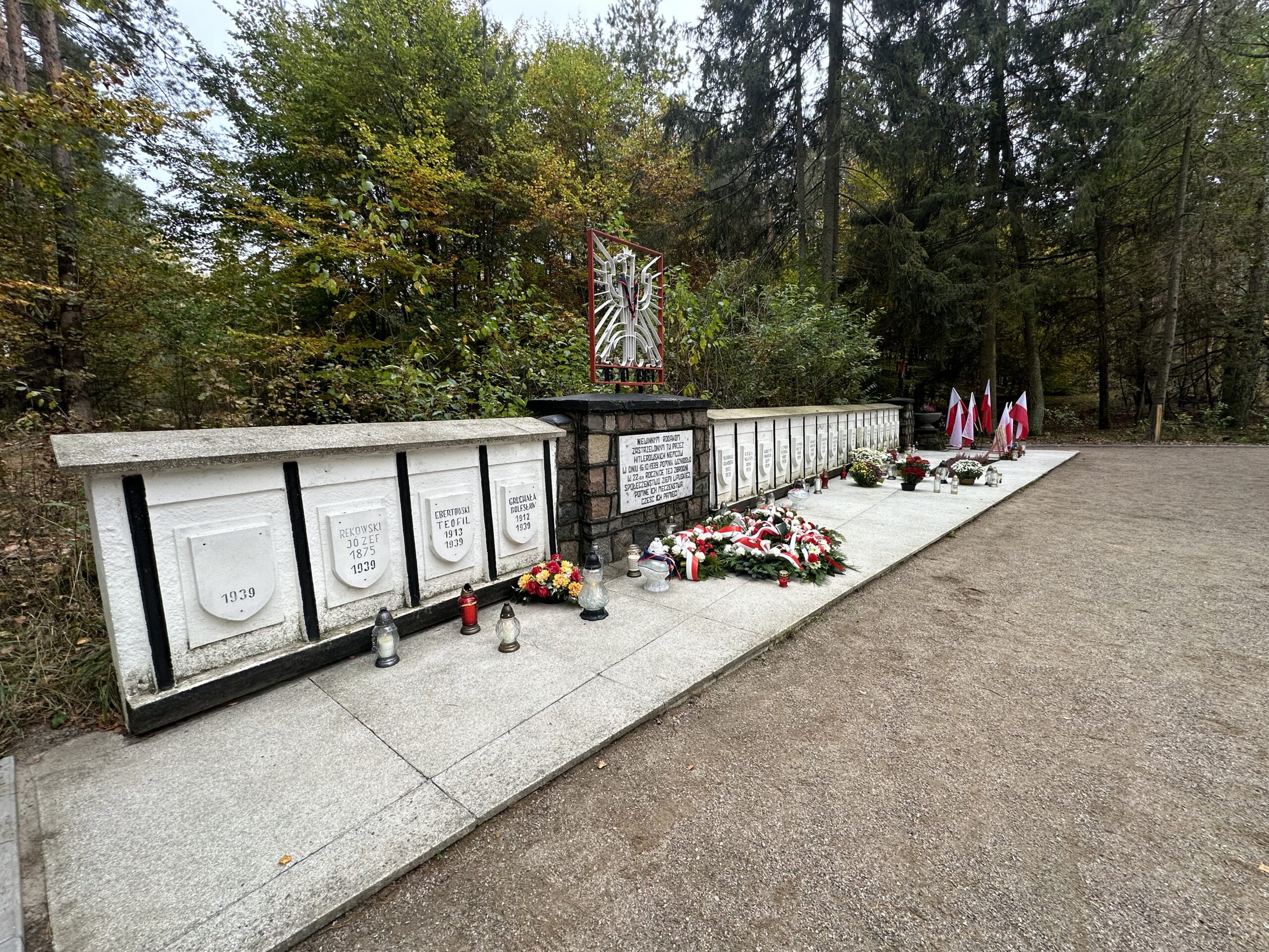 na zdjęciu widać groby pomordowanych w Karpnie na tle drzew. Z prawej strony znajduje się stojak z flagami biało-czerwonym. pomiędzy pomnikami znajduje siępamiątkowa tablica a nad nią orzeł polski wykonany z elementów metalowych w ramce w kształcie prostokąta